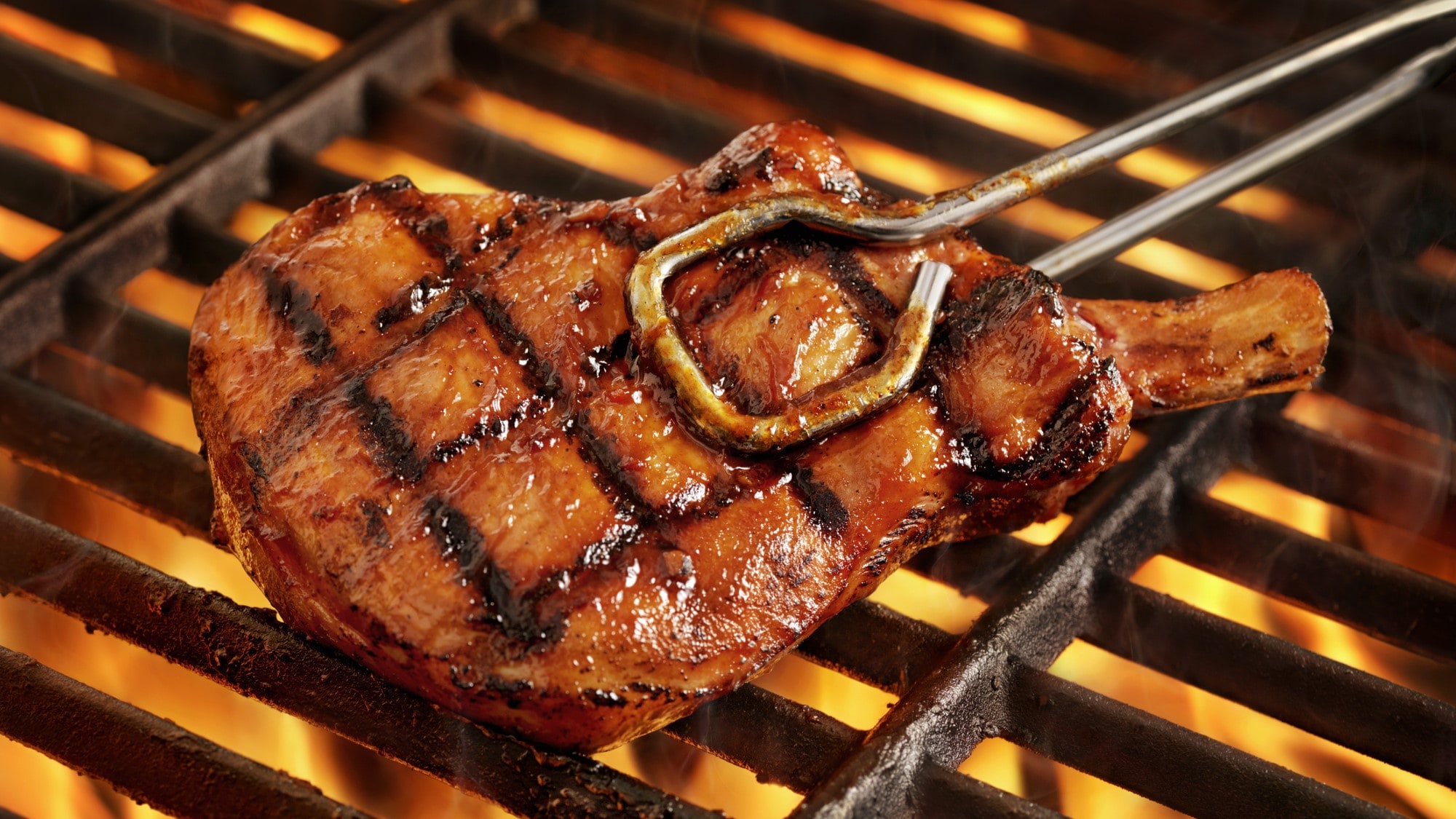 A pork chop on a hot grill with grill marks in both directions being held by a pair of tongs.